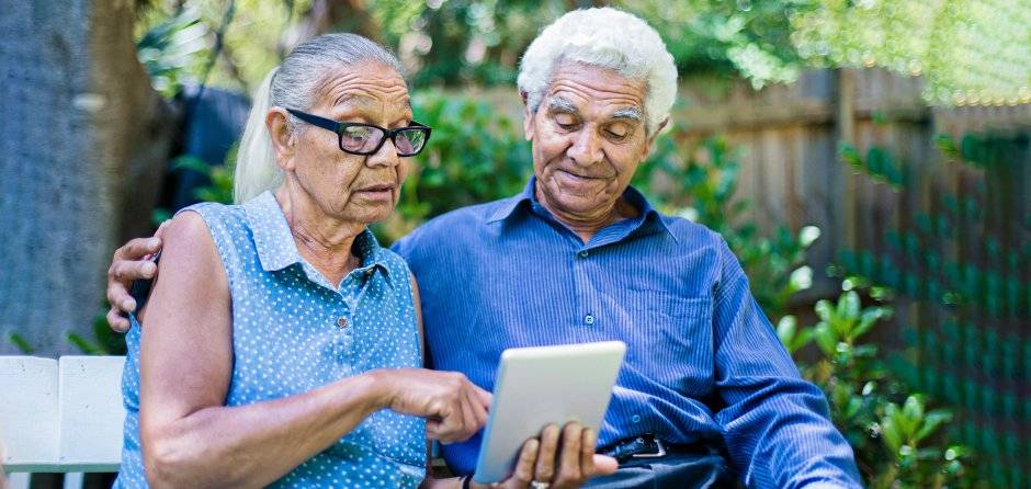 Older Aboriginal couple sitting on a bench outside looking at a smart tablet.