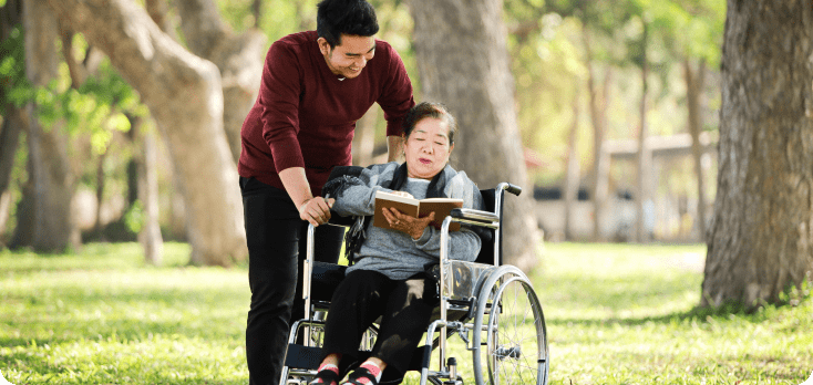 Young man with his grandmother who is in a wheelchair in the park, both reading a book.